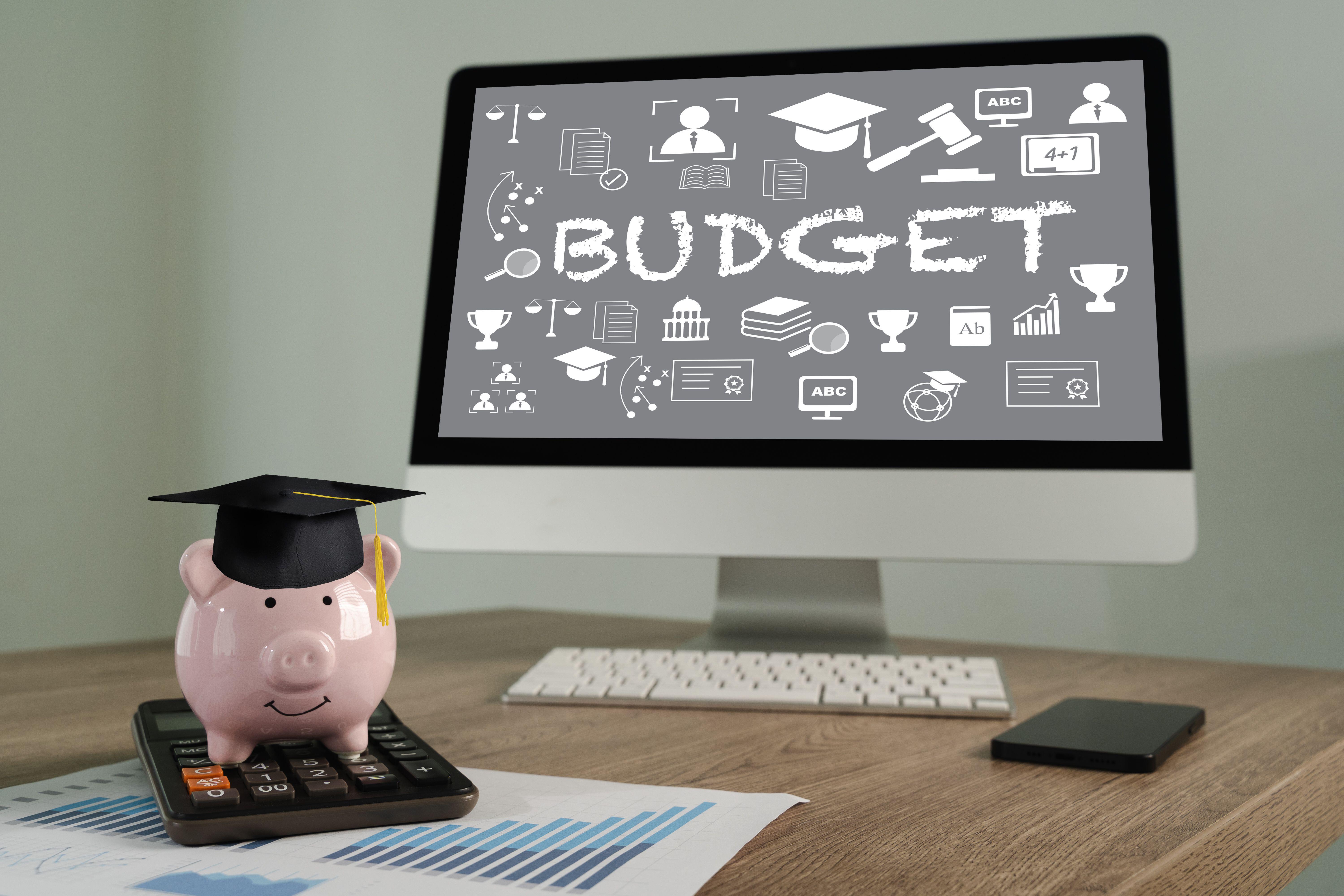 Student budget plan on computer monitor on a desk next to piggy bank wearing graduation cap and a calculator