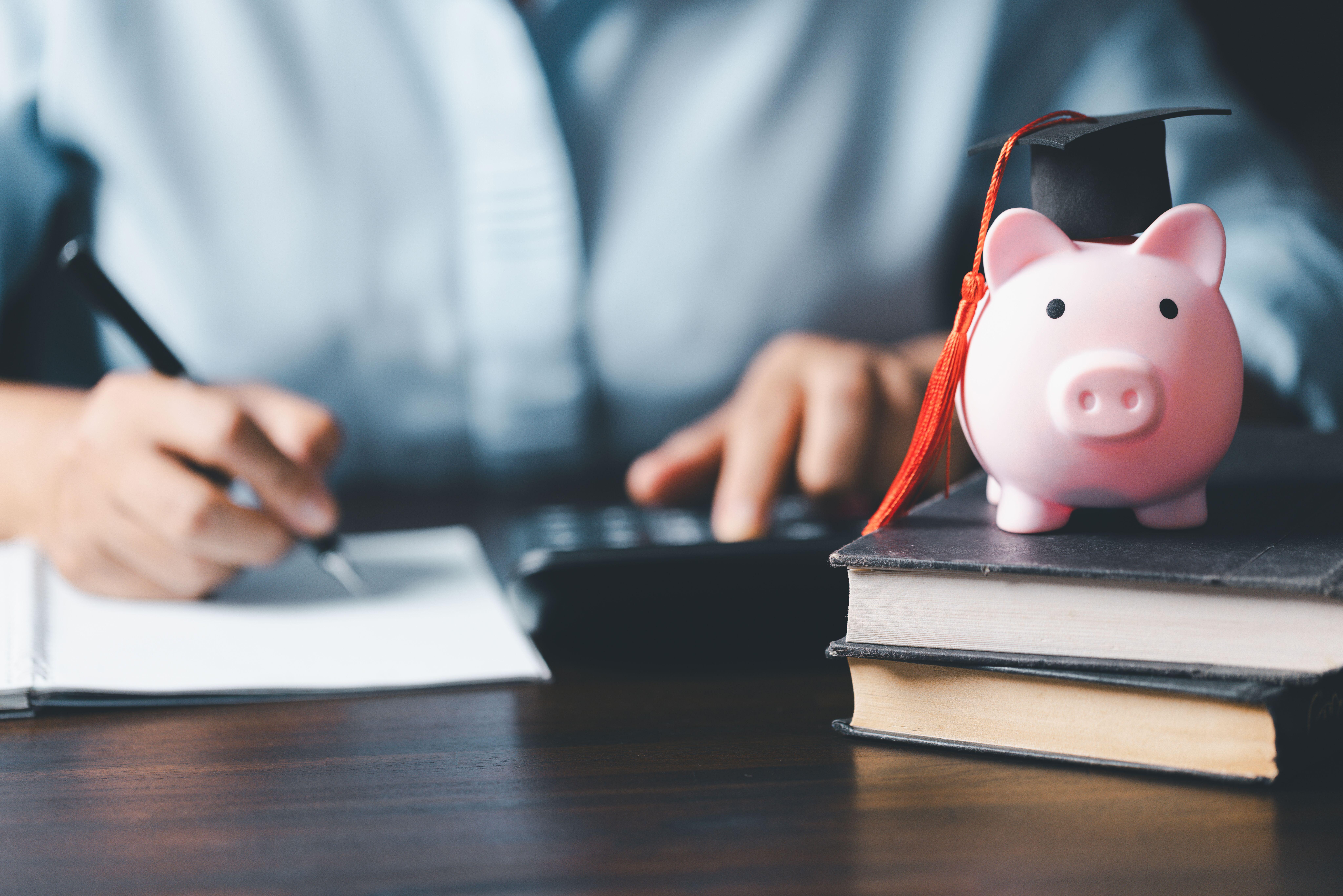 Man writing at desk next to piggy bank with graduation hat and pile of books