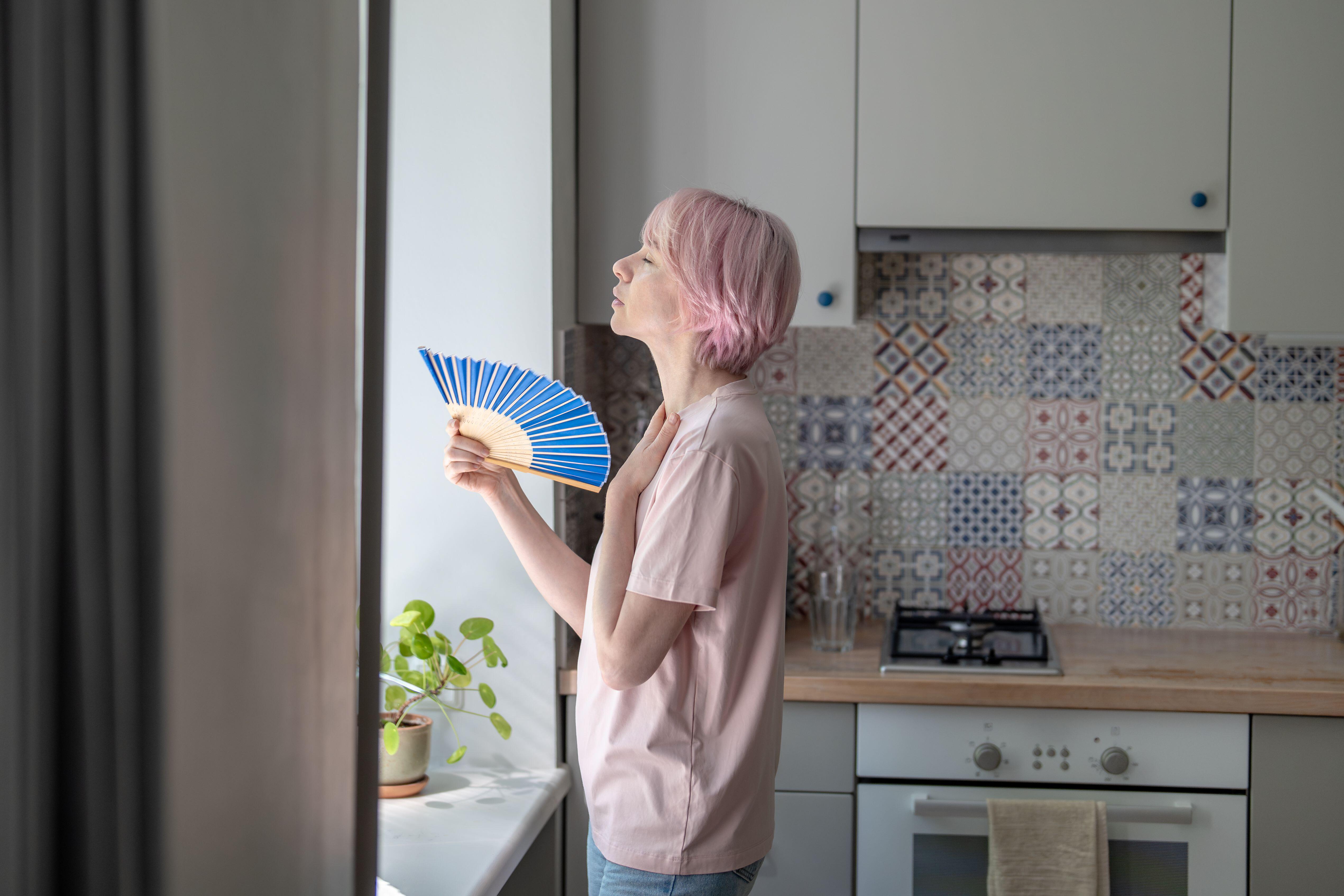 Hot, tired woman stood in kitchen using paper hand fan to cool down