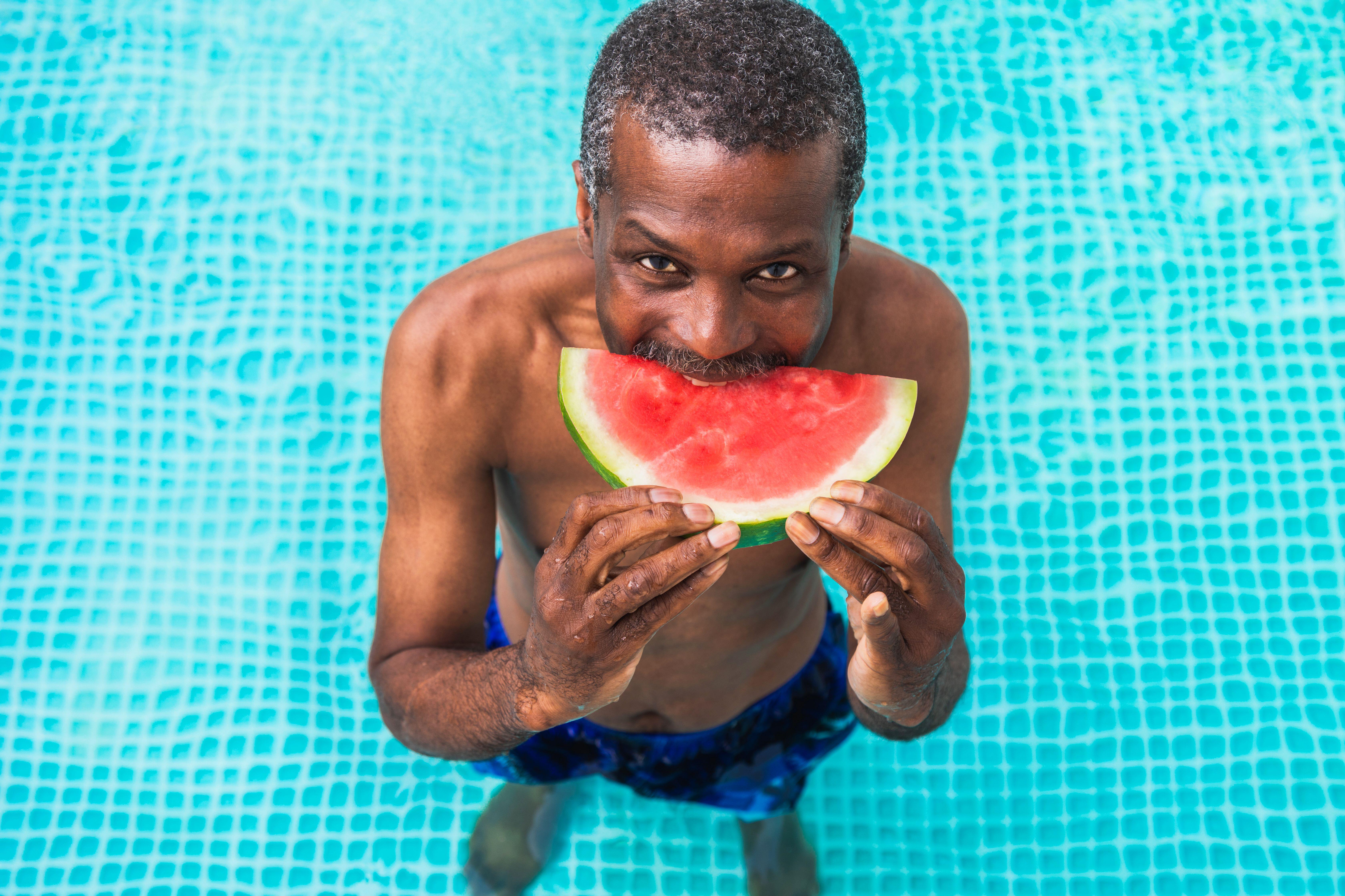 Happy senior man standing in a pool eating a slice of watermelon