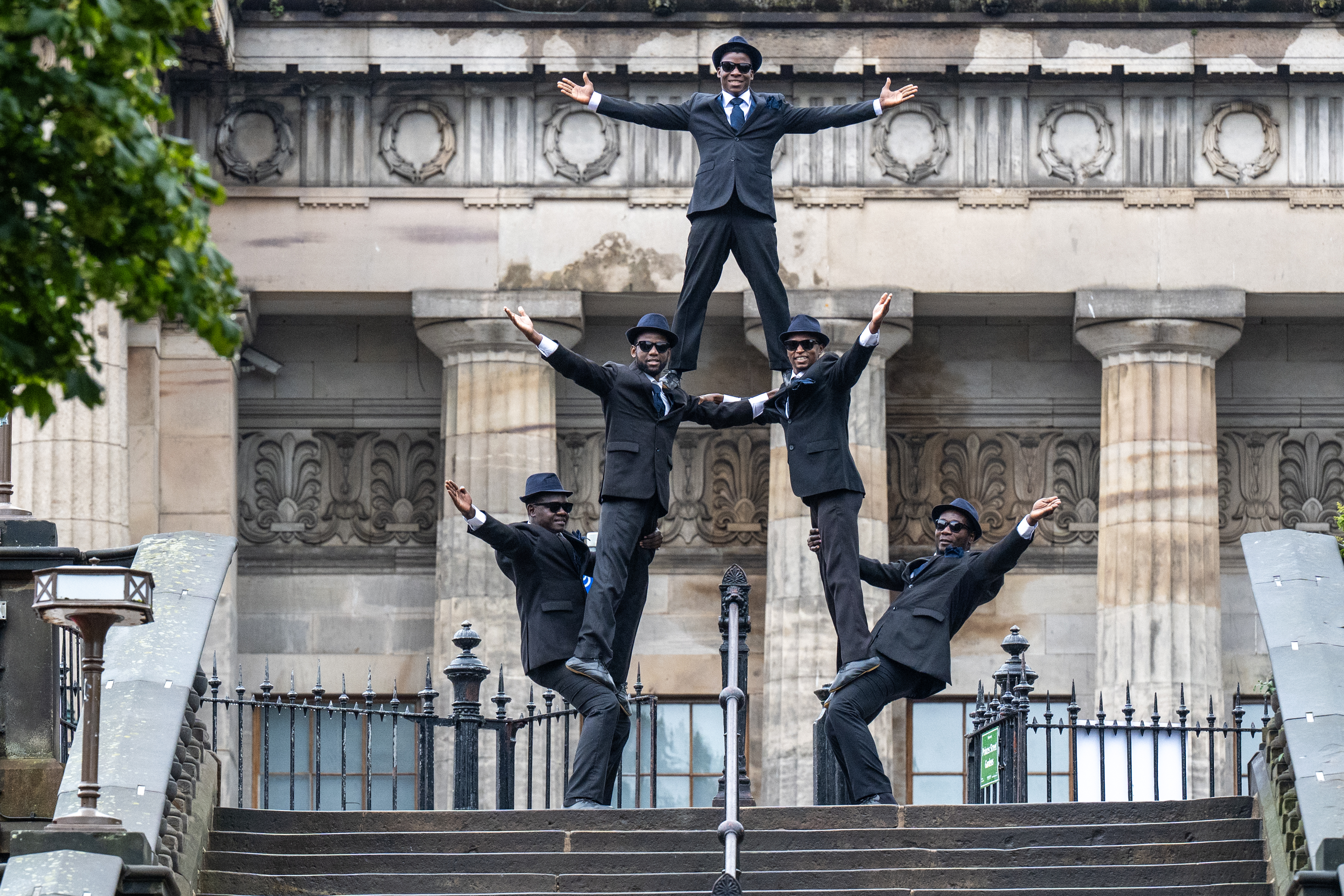 Performers wearing black suits and hats, standing on top of each other to form a pyramid