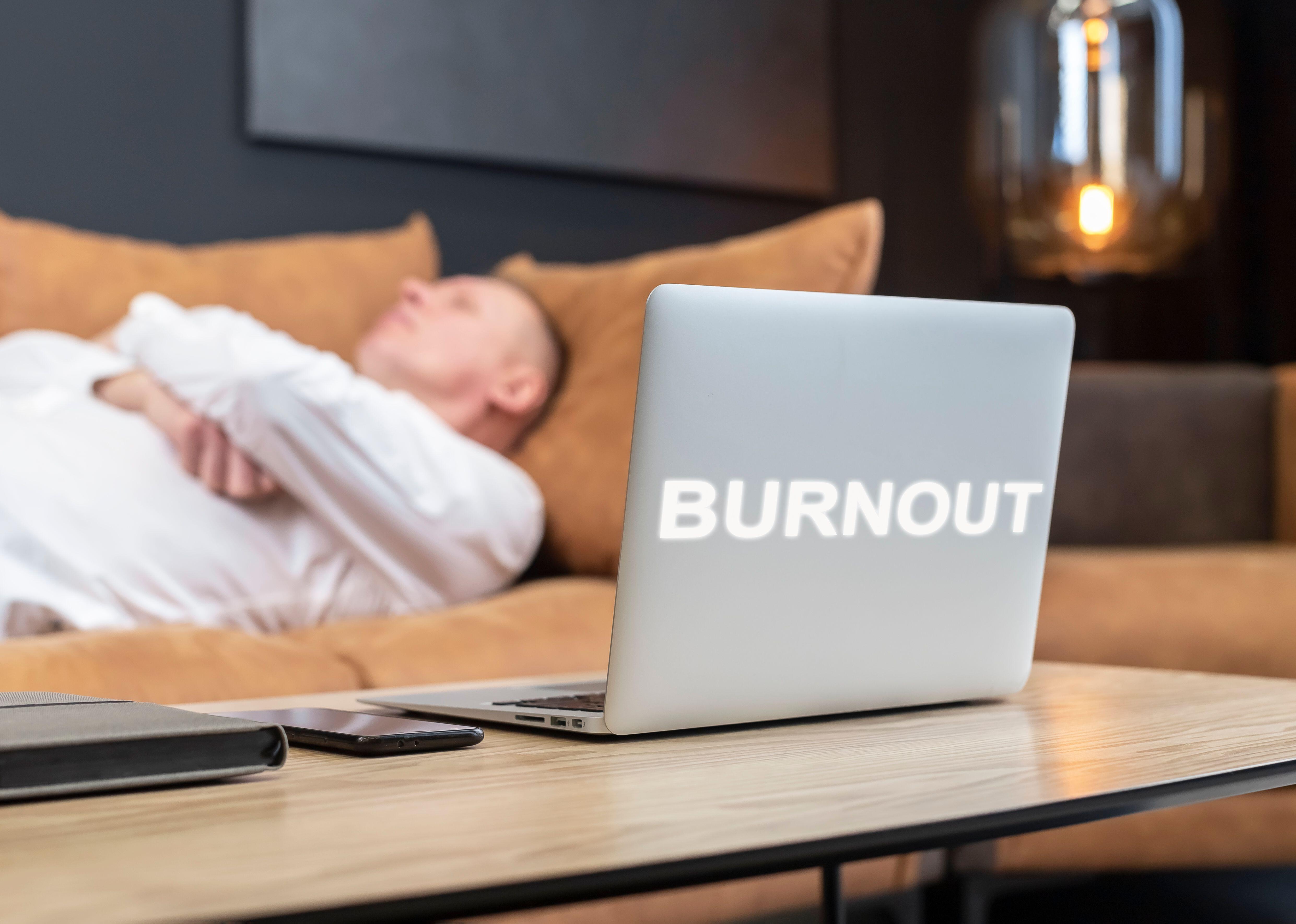 Exhausted businessman lying on sofa next to laptop which has burnout written on the cover