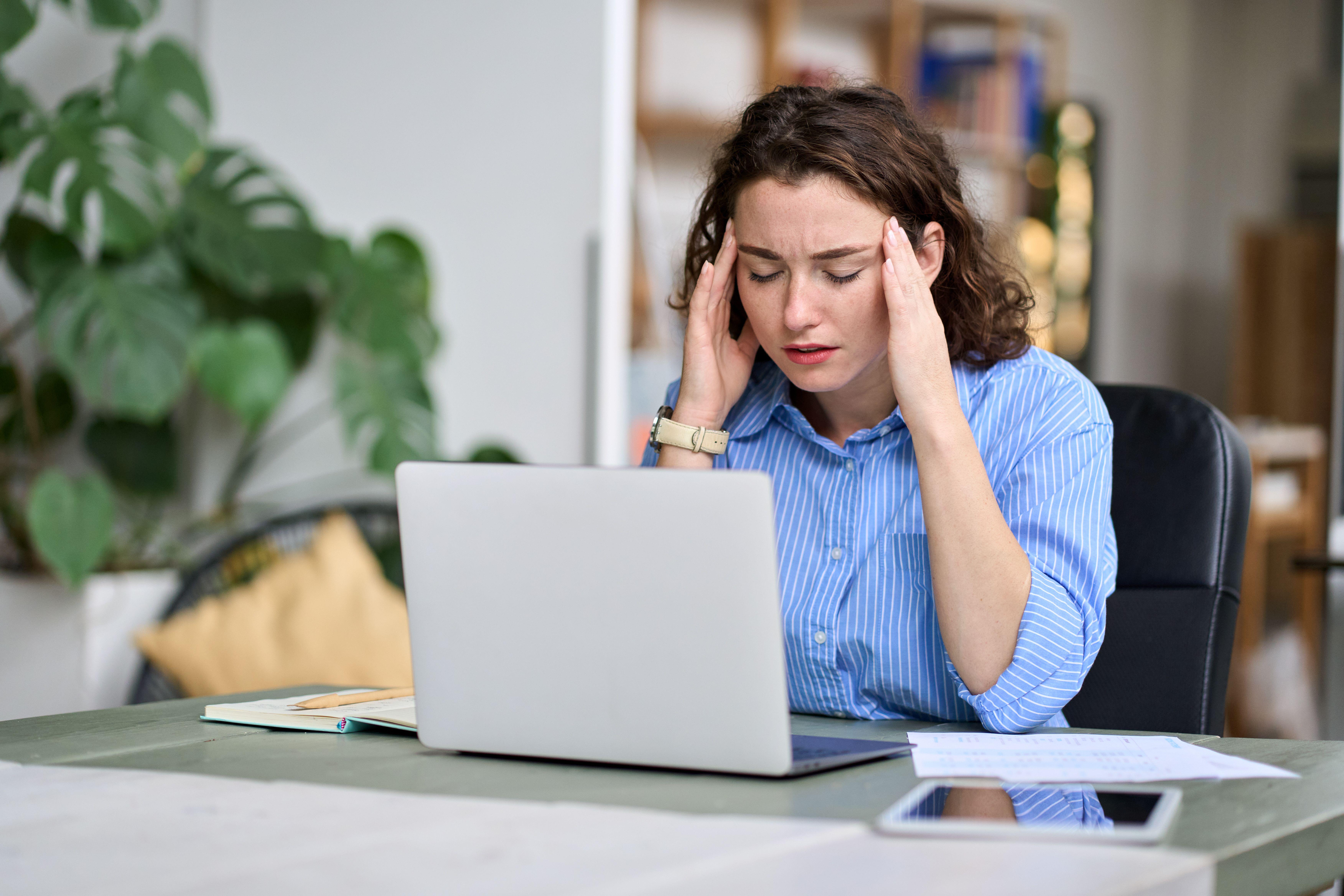 Stressed young woman sitting next to a laptop and holding her head in stress