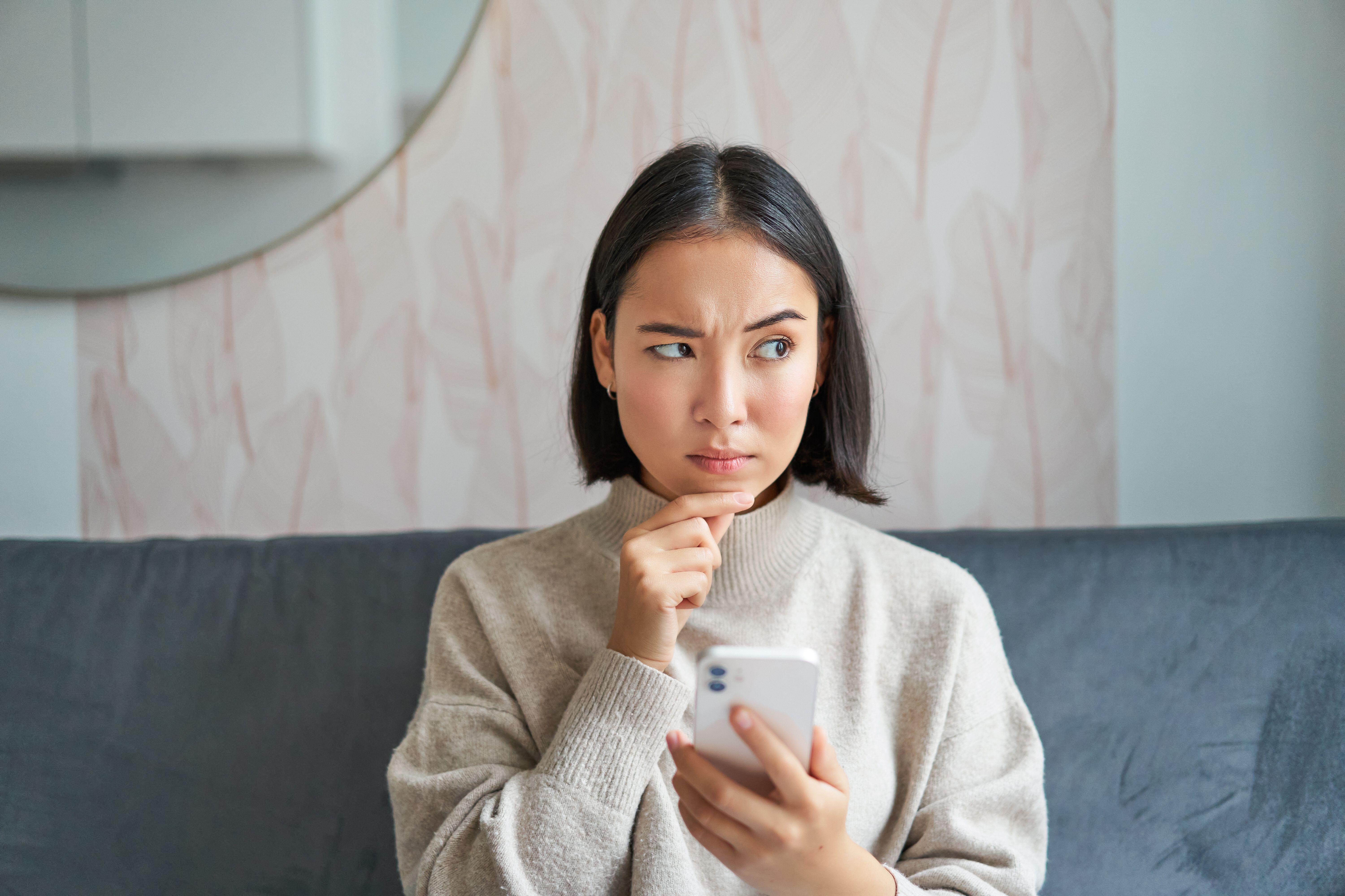 Portrait of girl sitting on sofa with smartphone, looking sceptical