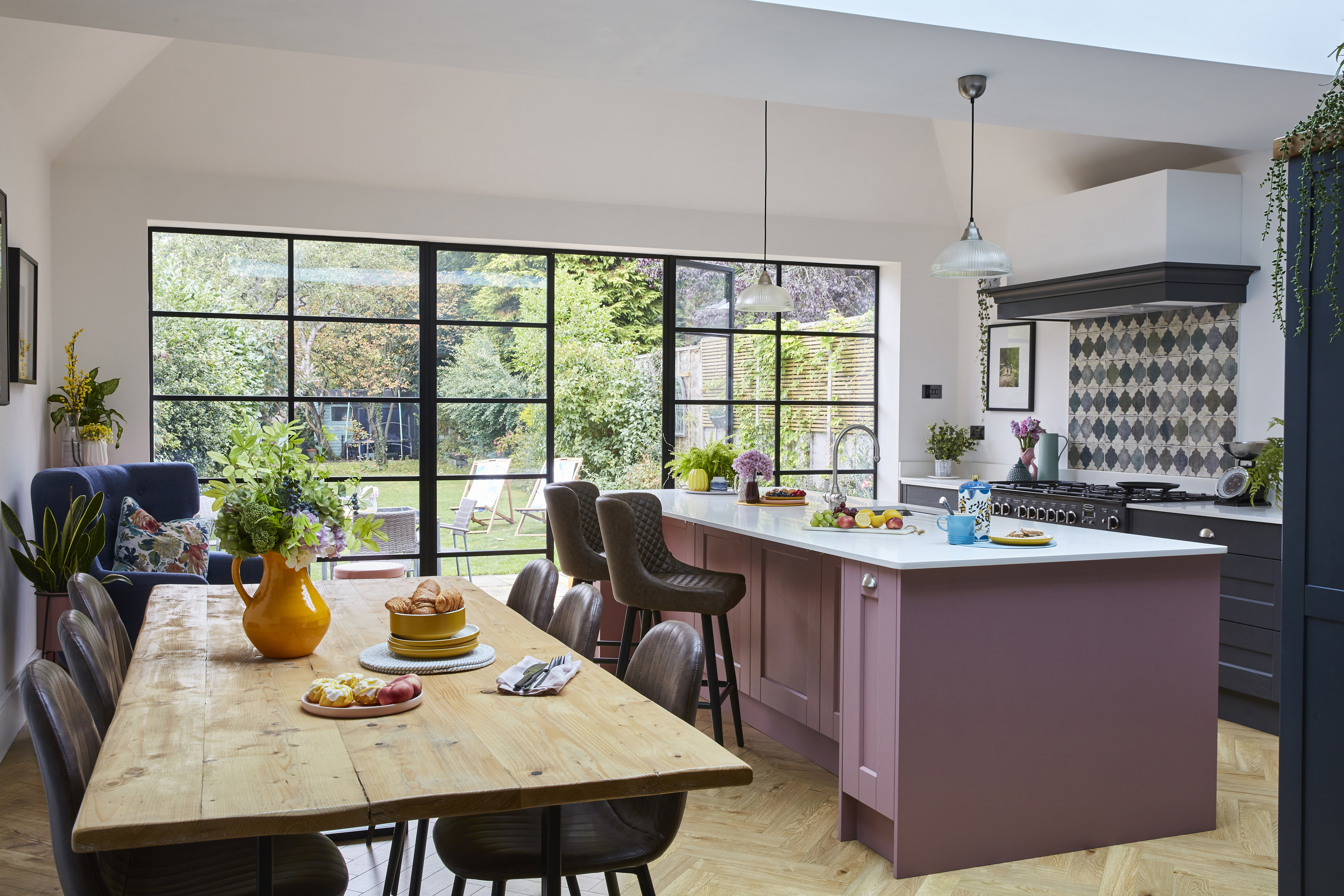 A colourful open-plan kitchen and dining area features a statement pink island and steel-framed glazing overlooking the garden