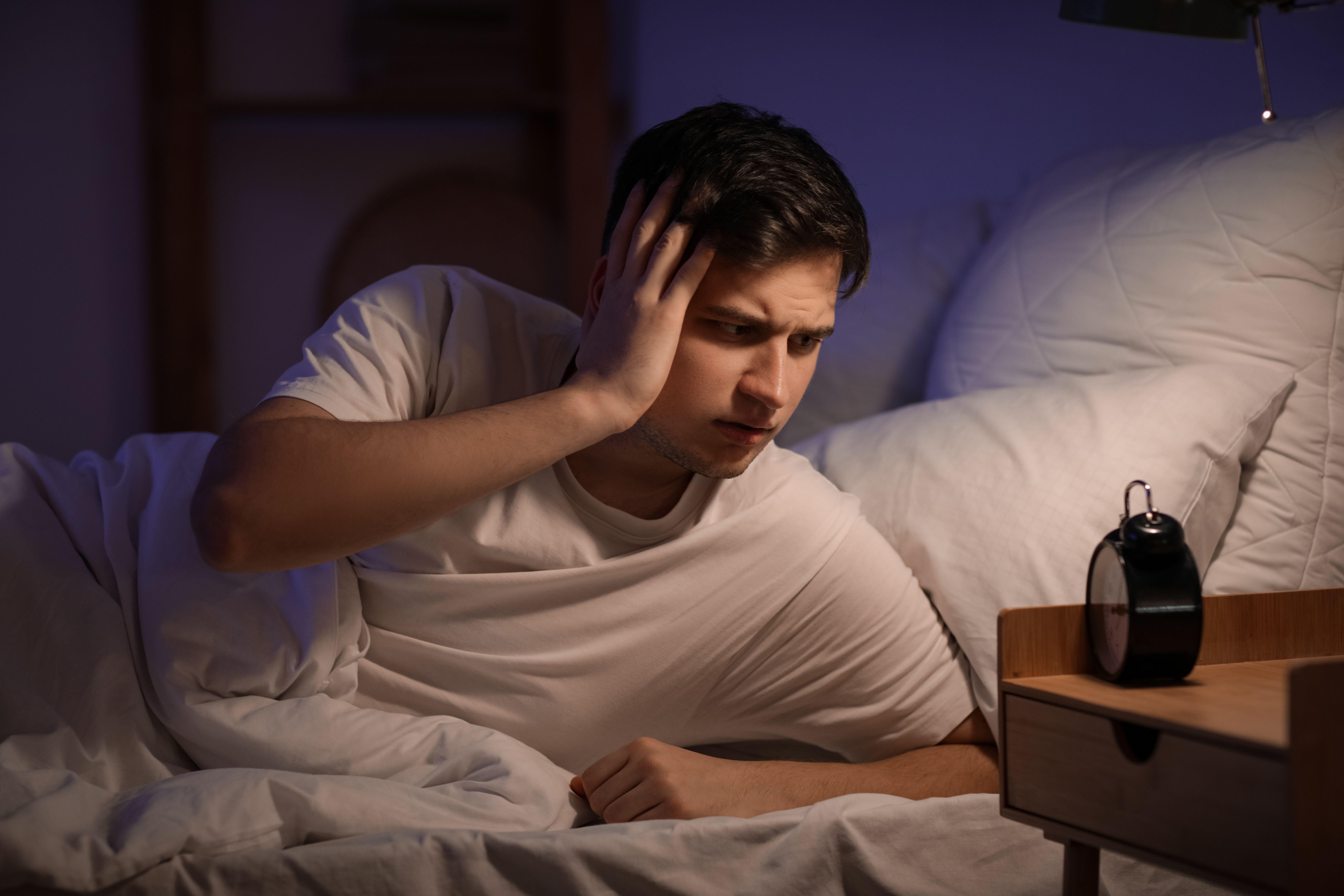 Young tired man waking up in dark looking at his alarm clock