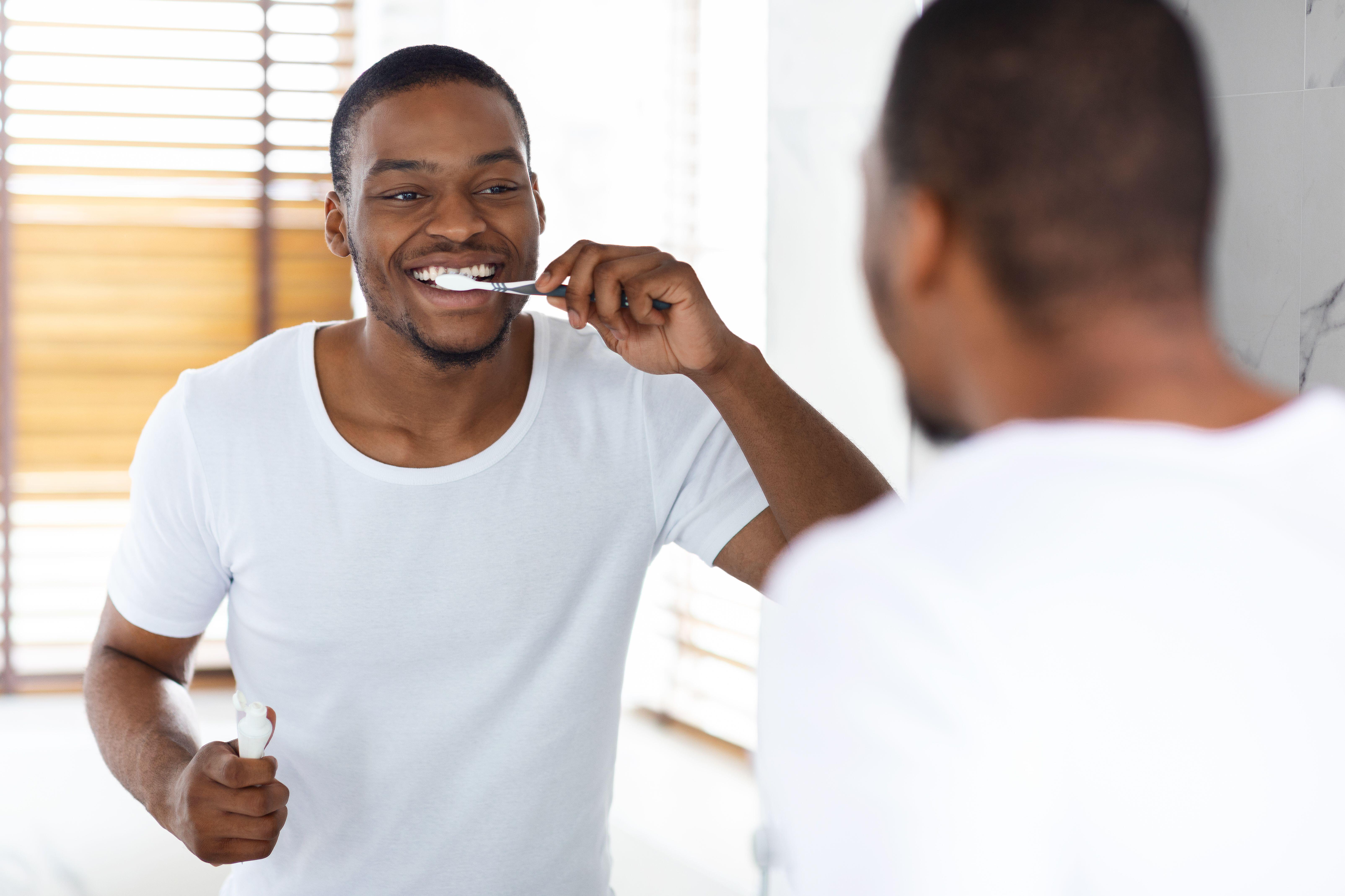 Black man brushing teeth while standing in front of mirror in bathroom at home