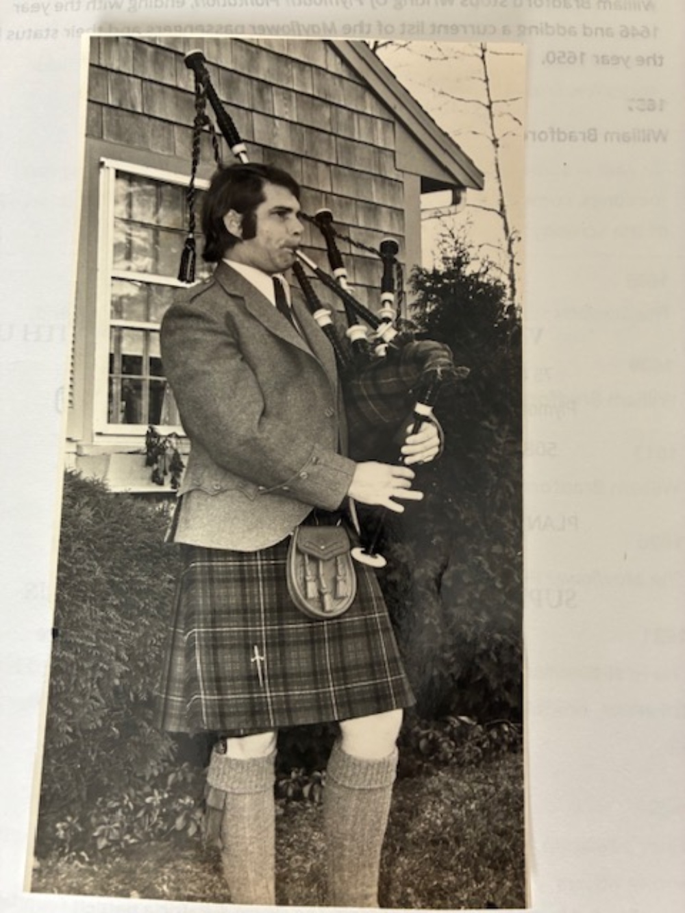 View of a black and white photo of Bruce Williams playing the bagpipes outside a house