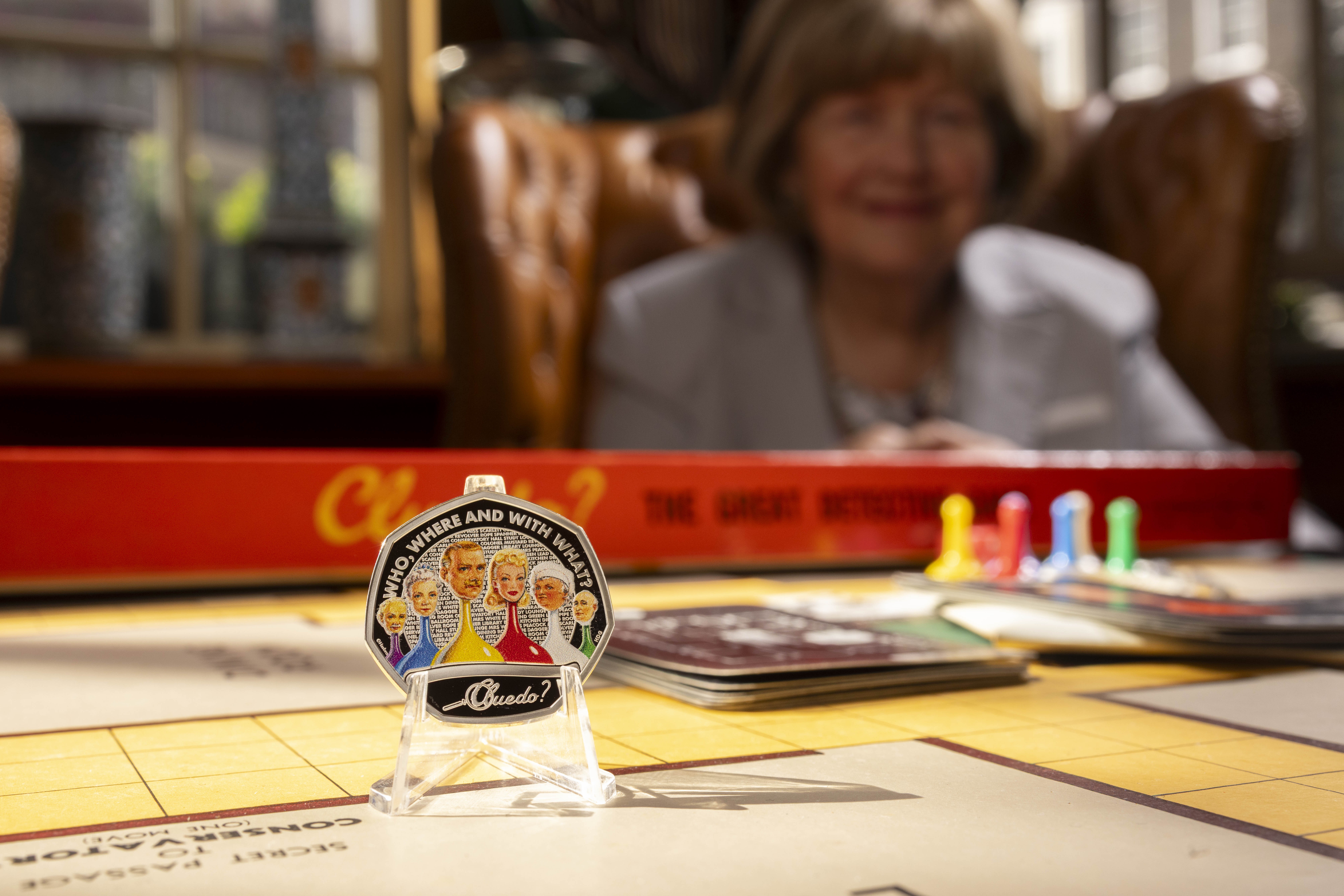 A Cluedo coin and game board with Marcia Lewis, daughter of the Cluedo creator, in the background