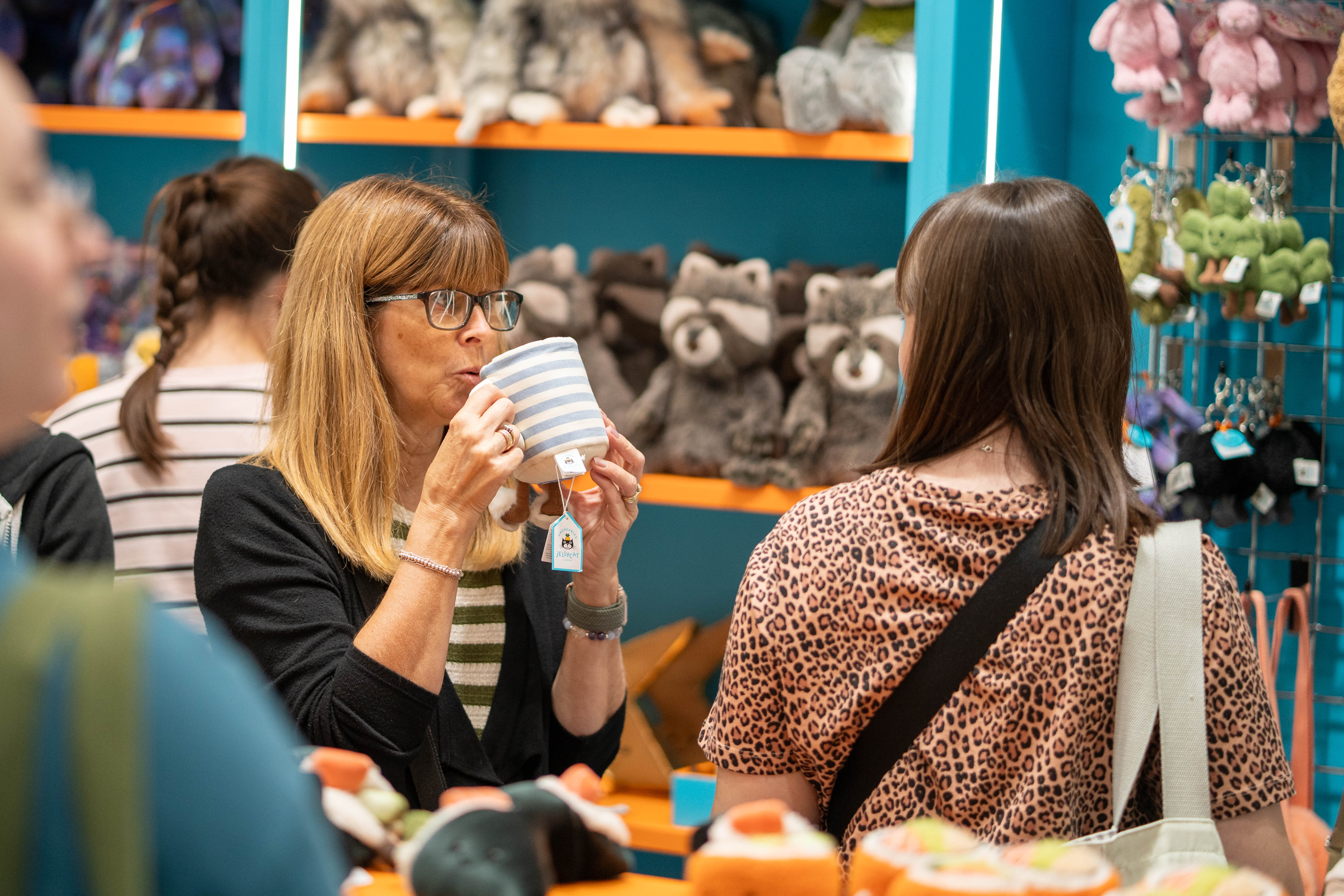 A woman pretending to drink from a cup-shaped Jellycat toy, in a toy shop