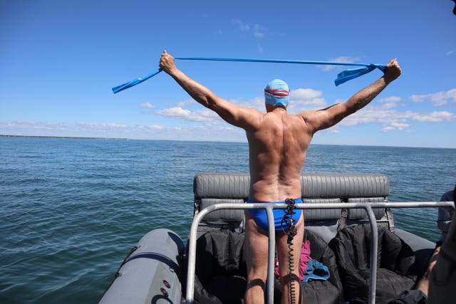 Lewis Pugh stands on a boat in Speedos, hat and goggles, stretching in front of a blue sky and calm sea