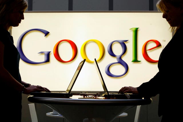 People work on laptops in front of an illuminated sign of the Google logo 