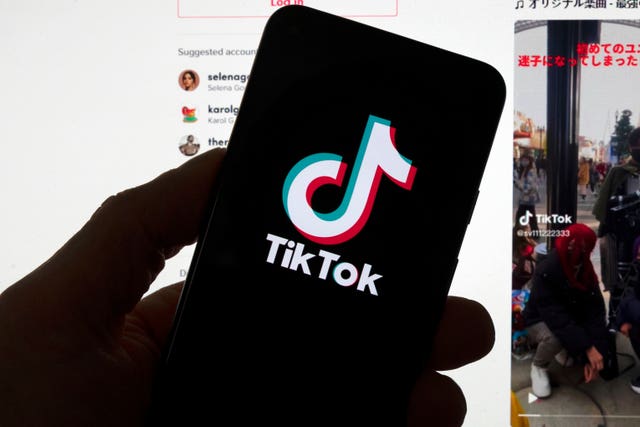 The TikTok logo is displayed on a mobile phone in front of a computer screen which displays a TikTok home page 