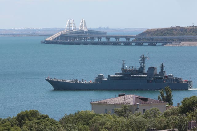 Russia military ship at sea just off a coast, with a large bridge in the background