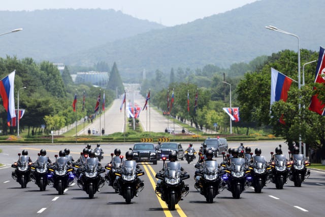 A motorcade of motorcycles with a car behind drives along a wide street 