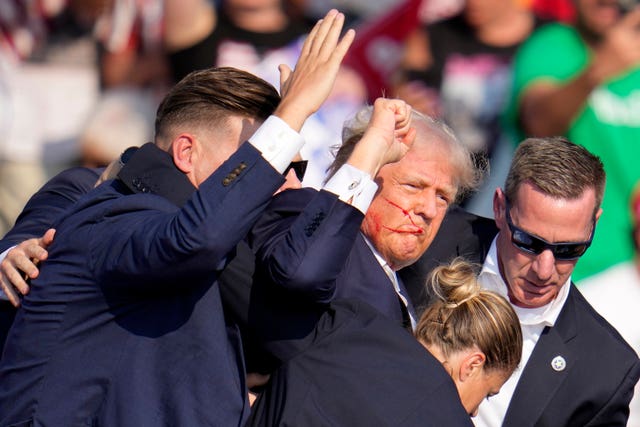 Trump, with blood across his face, holds his fist in the air as he's surrounded by secret service agents