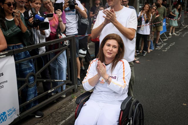 Christina Assi dressed in white top and trousers in a wheelchair at the Olympic torch relay in Vincennes with a man in a white T-shirt behind her and members of the crowd taking photographs