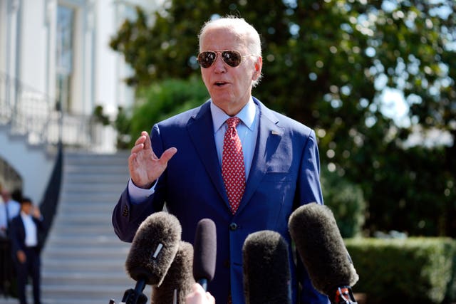 Joe Biden in sunglasses, with one had raised as he speaks in front of a number of microphones