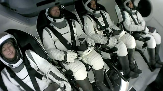 Four crew members shown strapped in their seats, wearing spacesuits