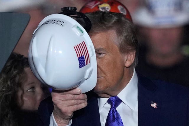 Donald Trump holding a white hard hat with a US flag in front of his face