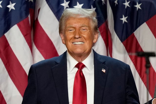 Donald Trump, in a suit and tie, smiles in front of an American flag