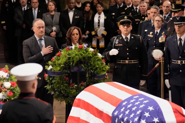 A coffin is covered in the US flag, Kamala Harris is visible over the top of a green wreath. Her husband stands with his hand over his heart.