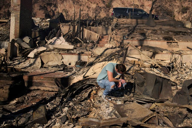 A man in a blue t-shirt, jeans and a mask kneels on the ground with his head down. Surrounding him as the charred remains of his father's home. Everything is burnt and ashen