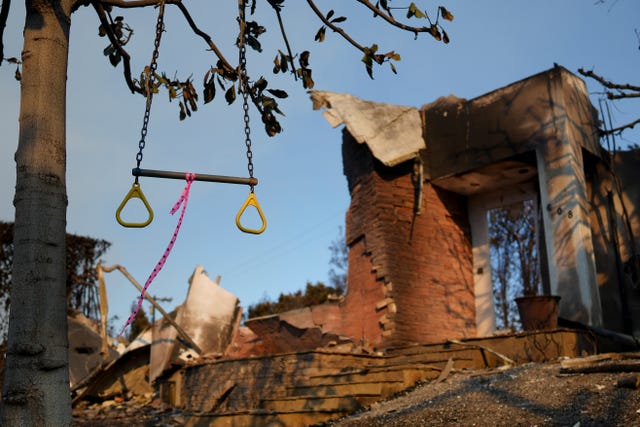 A rope swing with yellow handles and a pink rope, dangles from a tree. Behind it is the remnants of a house, what parts are still standing are blackened and burnt.