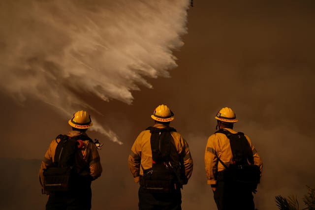 Three firefighters in yellow uniforms stand with their backs to the camera. They are looking as water is dumped on a scene in front of them. The details are hidden behind a brown, smoky sky