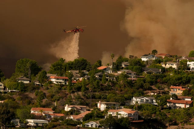 A red plane drops water on a landscape. There are houses and trees untouched by fire but the sky is dark and there is a huge plume of smoke in the distance