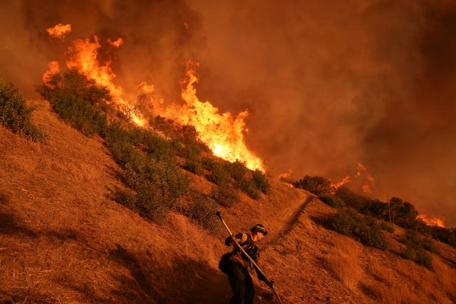 A firefighter holds a hose as they walk along a hillside. There are patches of grass that are on fire. Huge flames and lots of smoke are visible