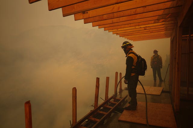 A firefighter stands with a hose and looks over the side of a building or a balcony, the air is filled with smoke and it is impossible to make out what he is looking at