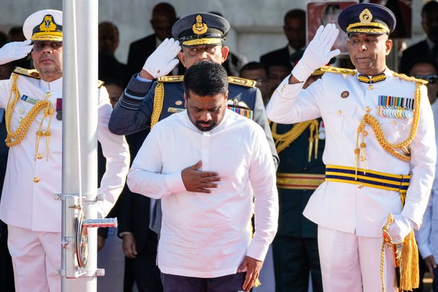 A man stands with his hand on his chest, behind him men in military dress salute