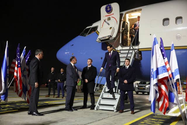 Marco Rubio on the steps of a plane, with dignitaries waiting to greet him on the tarmac 