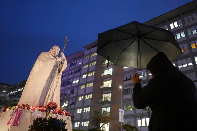 A man under an umbrella looks at a statue surrounded by flowers