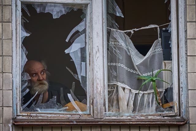 An elderly man looks out of his broken window 