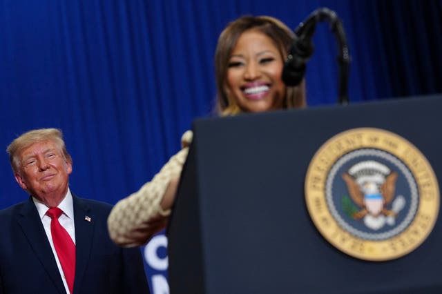 Donald Trump listens in the background as Nicki Minaj speaks from behind a lectern at a campaign rally